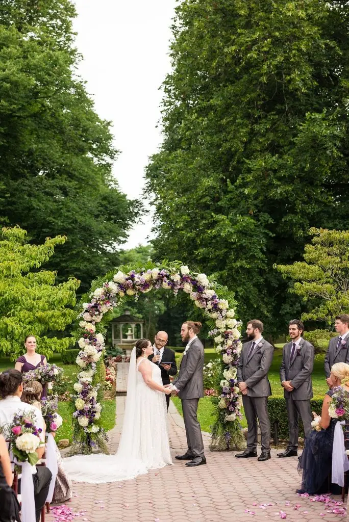 bride and groom beneath purple and white floral arch at their Mansion at Oyster Bay wedding outdoor ceremony - from Leah E. Moss Designs